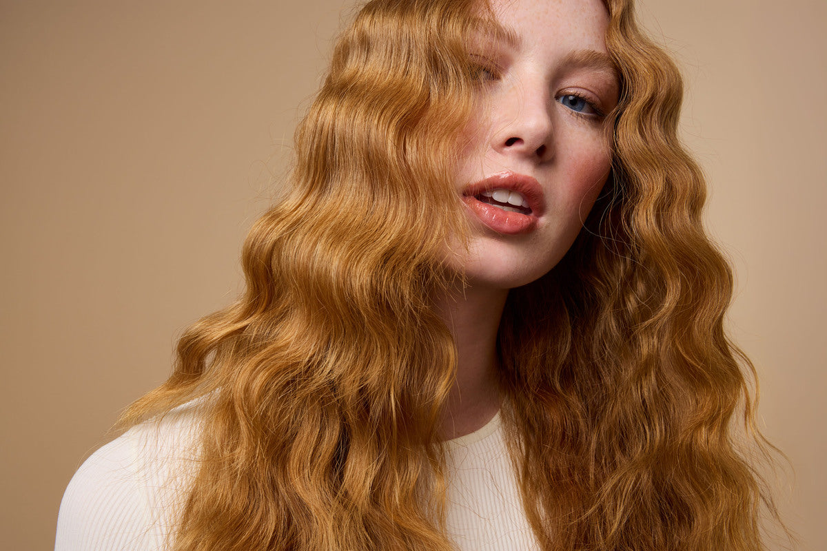 A person with long, wavy, reddish-blonde hair and fair skin poses against a beige background, wearing a light-colored ribbed top and looking slightly to the side with parted lips.