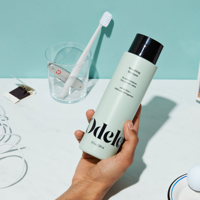 A hand holds a bottle of Odele Smoothing Shampoo over a white countertop, with a toothbrush in a clear cup, a matchbook, and tangled white earphones visible in the background.