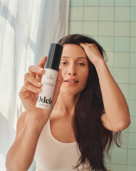 A woman with long dark hair holds up a bottle of Odele Dry Shampoo toward the camera, standing in front of a tiled wall, wearing a white tank top and touching her hair with her other hand.