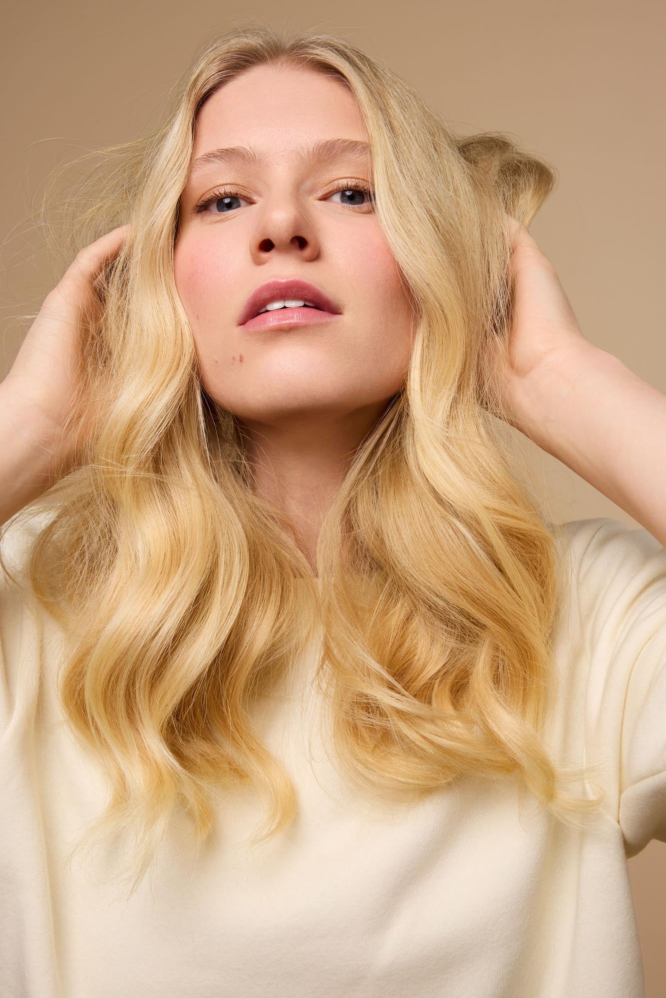 A woman with long, wavy blonde hair wearing a cream-colored top poses with her hands in her hair, looking confidently at the camera against a beige background.