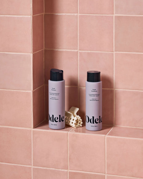 Two bottles of Odele hair product with black caps sit on a built-in shower shelf against pink tiles, next to a beige hair clip.