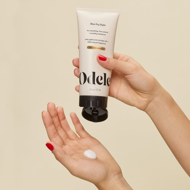 A person with red-painted nails dispenses a small amount of white Blow Dry Styler cream onto their palm, showcasing this hair styler’s heat protection benefits against a beige background.