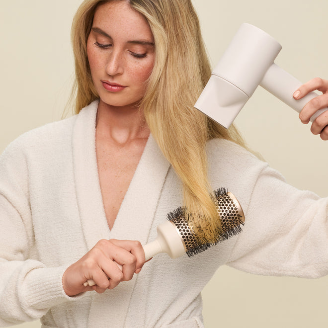 A woman in a white bathrobe uses the Blow Dry Styler, a white hair dryer, to blow-dry her long blonde hair while brushing it with a round brush for frizz control.