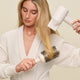 A woman in a white bathrobe uses the Blow Dry Styler, a white hair dryer, to blow-dry her long blonde hair while brushing it with a round brush for frizz control.