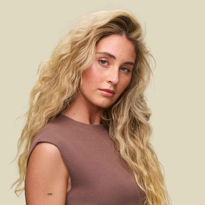A woman with long, wavy blonde hair and fair skin wears a sleeveless brown top against a beige background. Her smooth waves, defined with Curl Defining Foam, show flawless, humidity-resistant frizz control and a small "LOVE" tattoo on her arm.