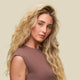 A woman with long, wavy blonde hair and fair skin wears a sleeveless brown top against a beige background. Her smooth waves, defined with Curl Defining Foam, show flawless, humidity-resistant frizz control and a small "LOVE" tattoo on her arm.