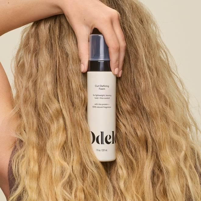 A person with long, wavy blonde hair holds Curl Defining Foam by their head, highlighting defined curls and the product’s silicone-free formula.
