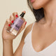 A woman in a white tank top sprays Hair + Body Mist on her neck, its sandalwood notes evoking a warm sauna. She wears red nail polish, with her partially visible face set against a light beige background.