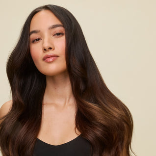 A woman with long, shiny hair and smooth skin poses against a light beige background, confidently showcasing frizz-free results achieved after heat styling with Thermal Protect Finishing Spray.