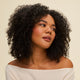 A woman with curly dark hair styled using Curl Defining Gel for frizz control and glossy finish looks to the side, wearing an off-the-shoulder cream top and natural makeup with blush, set against a plain light background.