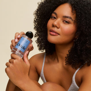 A woman with curly hair and glowing skin holds a bottle of Hair + Body Mist by Odele near her face, wearing a gray tank top against a neutral background, evoking warm notes of amber and vanilla.