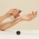 Wearing red nail polish, a person spritzes Hair + Body Mist from a clear bottle onto their wrist; the bottle, labeled Ode and featuring bergamot leaf notes, has its black cap set on the white surface below.