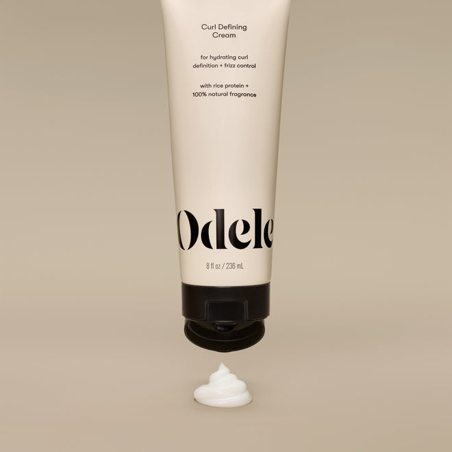 A beige tube of Curl Defining Cream stands upside down with a swirl of white cream below its black cap, set against a neutral background.