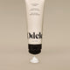 A beige tube of Curl Defining Cream stands upside down with a swirl of white cream below its black cap, set against a neutral background.