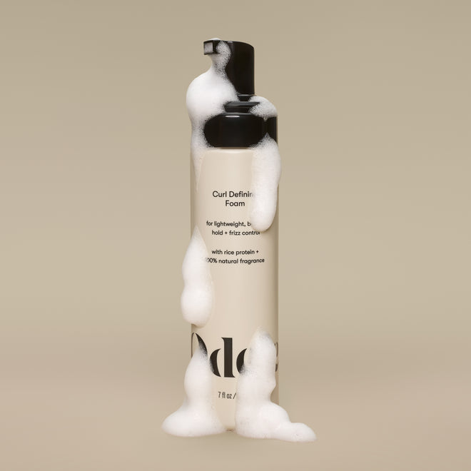 A bottle of Curl Defining Foam, covered in white suds against a beige background, features a label highlighting rice protein, natural fragrance, and its silicone-free, humidity-resistant frizz control benefits.