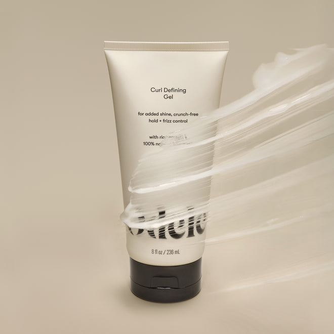A tube of Curl Defining Gel stands upright on a beige surface, partially covered by a glossy smear. The minimalistic, clean setting highlights the product's frizz control benefits.