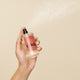 A hand sprays a clear bottle labeled “Odele Hair + Body Mist” against a beige background, releasing a fine bergamot leaf-infused mist from the nozzle.