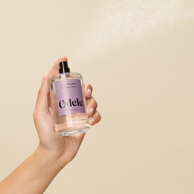 A hand sprays Odele Hair + Body Mist, featuring a light purple label, against a beige background. A fine sandalwood-infused mist is visible in the air.