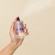 A hand sprays Odele Hair + Body Mist, featuring a light purple label, against a beige background. A fine sandalwood-infused mist is visible in the air.