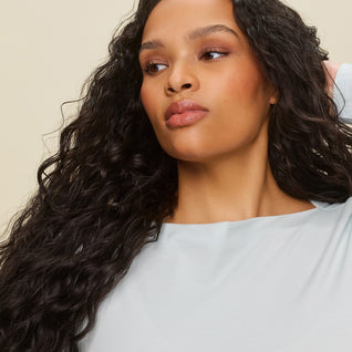 A person with long, wavy dark hair styled with Curl Defining Cream poses in a light blue top, one hand behind their head, looking off to the side against a plain light background.