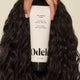 A hand holds a tube of Curl Defining Cream with wavy, dark hair in the background. The label highlights its hydrating formula for curl definition, frizz control, rice protein, and natural fragrance.