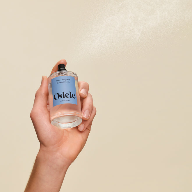 A hand holds a bottle of Hair + Body Mist against a beige background, releasing a fine spray scented with warm amber and vanilla into the air.