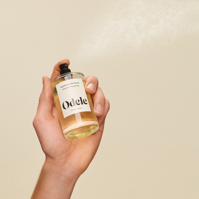 A hand sprays a fine mist of Signature Hair Scent from a clear bottle against a neutral beige background.