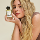 A woman with long, wavy blonde hair holds a bottle of Signature Hair Scent near her face, gazing softly at the camera. The scent features notes of cucumber and bamboo, set against a neutral background.