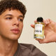 A person with short curly hair holds a bottle of Signature Hair Scent near their face, highlighting its label. The plain beige background reflects the fresh, crisp notes of cucumber and bamboo found in the scent.