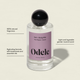 A clear glass bottle of Odele Hair + Body Mist, featuring a black cap and pink label. Showcases its natural fragrance, hydrating botanicals, and a light, gender-neutral sandalwood scent ideal for layering.