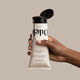 A hand holds an upside-down tube of Odele Air Dry Styler against a plain, light-colored backdrop. Product drips down the front of the bottle as if it was recently squeezed.