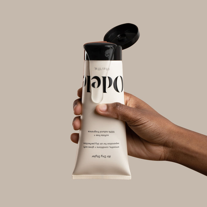 A hand holds an upside-down beige Air Dry Styler Mini with a black flip-top cap and bold black text, making it stand out as your go-to anti-frizz styling cream against the plain background.