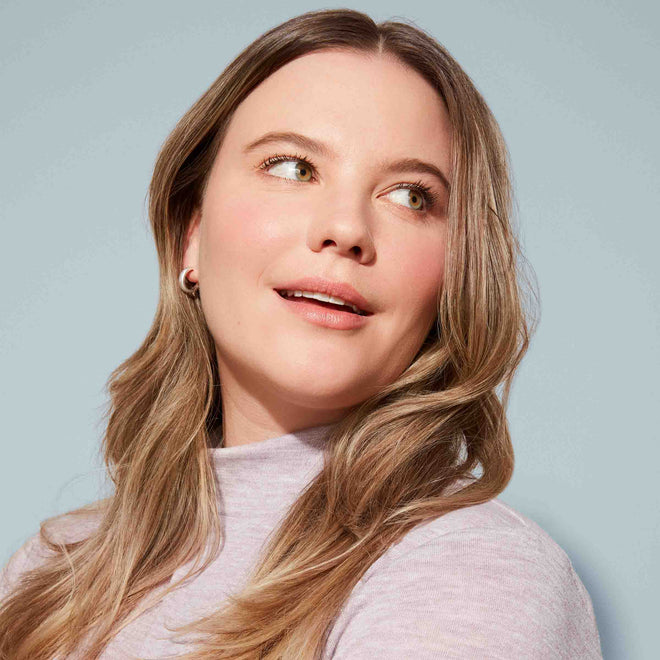 A woman with long, wavy light brown hair and fair skin smiles softly, her smooth anti-frizz locks styled by the Air Dry Styler Mini. Wearing a light purple top and small hoops, she poses against a light blue background.
