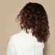 A young woman wearing a white button-up shirt stands against a light-colored background with her back turned to the camera to showcase her medium-brown mid-length curls.