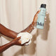 A person holds a foaming white shower pouf and a bottle of Odele Clarifying Body Wash, lather covering their arms against a beige shower curtain background.