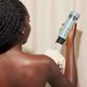 A woman with medium-dark skin and braided hair stands with her back to the camera as she squeezes Odele Clarifying Body Wash onto a white shower pouf.