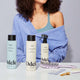 A woman with curly hair sits behind a white tabletop where Odele Clarifying Shampoo, Odele Curl Defining Conditioner and Odele Leave-in Detangling Tonic sitsamong various beauty items. She wears a blue tank top and off-shoulder sweater.