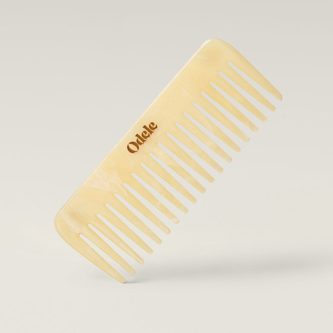 A light beige wide-toothed comb with a gold Odele logo on the handle stands at an angle against a plain, light-colored background.