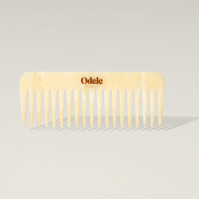 A light beige wide-toothed comb with a gold Odele logo on the handle stands upright against a plain, light-colored background.