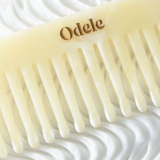 A light beige wide-toothed comb with a gold Odele logo on the handle lays on a background of swirling white cream.