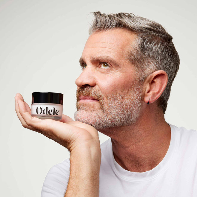 A middle-aged man with gray hair and a beard holds a small jar labeled Odele Beauty Cream Pomade near his face, looking upward thoughtfully in front of a plain light-colored background. He wears a white shirt and has a small hoop earring in his left ear.