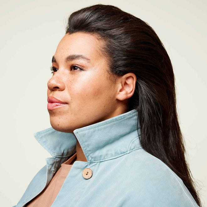 A young woman with light brown skin, freckles, and long dark hair looks to the side in front of a plain light-colored background.