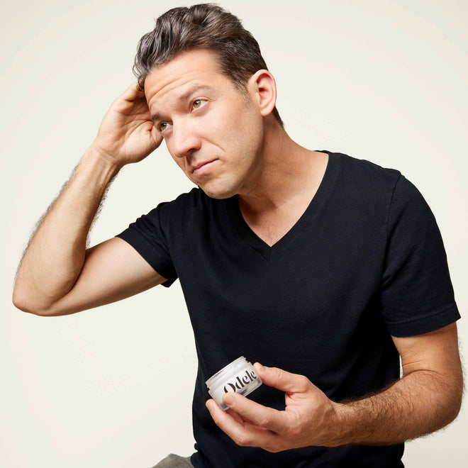 A clean-shaven man with pale skin and grey hair wears a black V-neck t-shirt while styling his hair with the Odele Cream Pomade in front of a plain light-colored background.