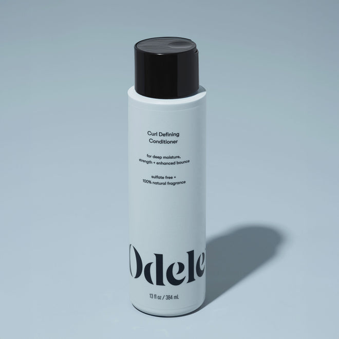 A bottle of Odele Curl Defining Conditioner stands upright against a plain, light blue backdrop with the front of the label facing the camera.