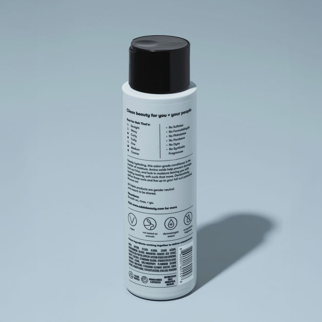 A bottle of Odele Curl Defining Conditioner stands upright against a plain, light blue backdrop with the back of the label facing the camera.