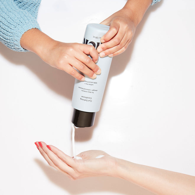 The hands of a young child squeeze a bottle of Odele Curl Defining Conditioner into the outstretched hand of an adult.
