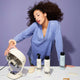 A young woman with light brown skin and curly hair wears a light blue top while she leans over a white table with an assortment of Odele products and other beauty accessories.