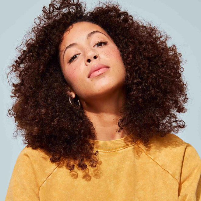 A young woman with curly brown hair wears a yellow sweatshirt and hoop earrings while confidently posing in front of a plain, light blue background.