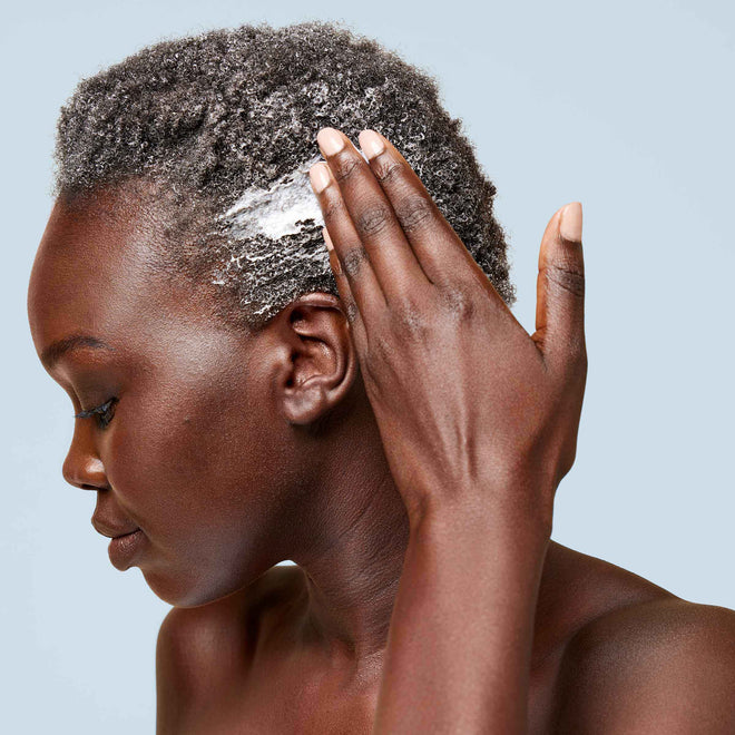 A young woman with dark brown skin and short coily hair swipes a handful of the Odele Curl Defining Conditioner onto the side of her head in front of a plain, light-colored background.