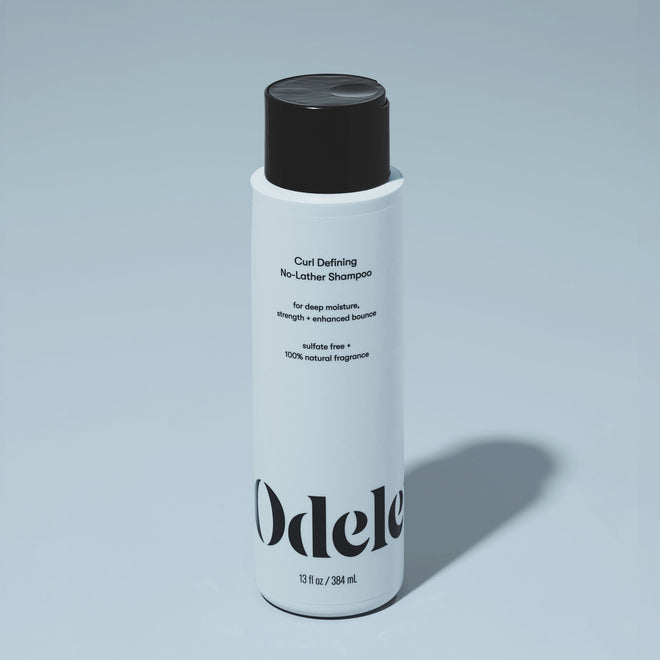 A bottle of Odele Curl Defining No-Lather Shampoo stands upright against a plain, light blue backdrop with the front of the label facing the camera.
